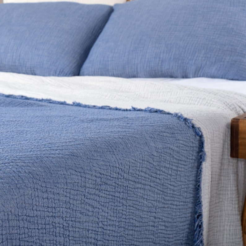 Puede incluir: Una manta de tiro acolchada azul con un borde con flecos. La manta est&aacute; doblada en una cama con almohadas blancas y azules.