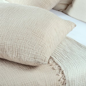 May include: Close-up of a bed with beige bedding. The image features a textured, beige pillow and a matching blanket with a fringed edge. Additional pillows are visible in the background, all in a neutral color palette.