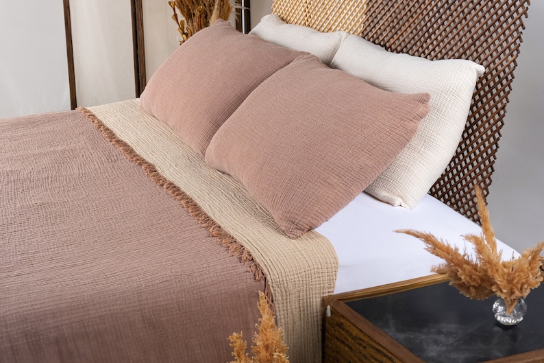 Puede incluir: Una manta de tiro marr&oacute;n y beige con un borde de flecos en una cama con una cabecera blanca y detalles de mimbre marr&oacute;n. La cama est&aacute; hecha con s&aacute;banas blancas y dos almohadas marrones.