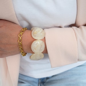 Peut inclure: Un bracelet à maillons doré et un bracelet épais crème avec de grosses perles rondes sont portés au poignet. La personne porte une chemise blanche et un blazer rose clair.