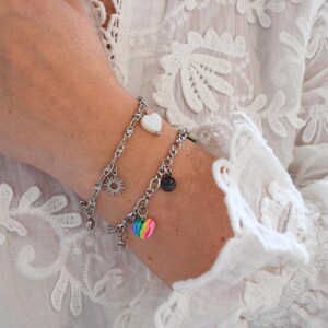 Peut inclure: Deux bracelets à breloques argentés avec divers charms, dont un cœur blanc, un soleil, un cœur arc-en-ciel et un smiley. Les bracelets sont portés sur un poignet, sur un fond de dentelle blanche.