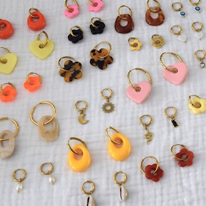 Può includere: Una collezione di orecchini con cerchi dorati e vari ciondoli. I ciondoli includono cuori, fiori e forme astratte in colori come giallo, arancione, rosa e marrone. Alcuni orecchini presentano piccoli ciondoli pendenti.