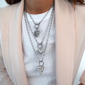Peut inclure: Colliers argentés superposés avec trois pendentifs différents : un soleil, un cœur et une flèche texturée. Les colliers sont portés sur un t-shirt blanc et un blazer rose clair. Les pendentifs sont fixés aux chaînes par des fermoirs circulaires.