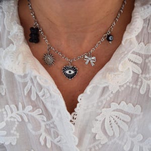 Peut inclure: Un collier argenté orné de plusieurs breloques : un ours en gélatine noir, un soleil, un cœur avec un œil, un nœud et une boule 8. Le collier est porté sur un chemisier blanc en dentelle.