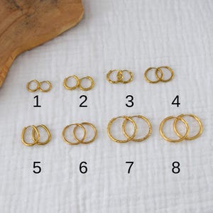 Peut inclure: Présentation de huit paires de boucles d'oreilles créoles dorées, chacune avec un design unique. Les boucles d'oreilles sont disposées sur un tissu blanc, numérotées de 1 à 8. Différentes tailles et textures sont proposées.