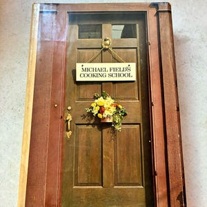 Puede incluir: Portada de libro de cocina que presenta una puerta de madera con un letrero que dice "MICHAEL FIELD'S COOKING SCHOOL". La puerta está adornada con un arreglo floral. La portada tiene un marco marrón y rojo.