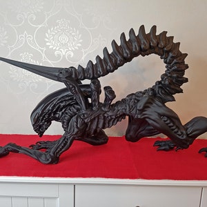 Op de afbeelding: Een zwart 3D-geprint alien-wezentje met een lange staart en scherpe tanden. Het wezen zit in een hurkende positie met zijn hoofd naar beneden en zijn armen uitgestrekt.