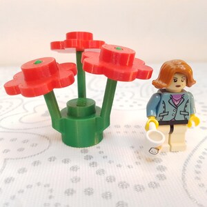 Könnte beinhalten: Ein Lego-Set mit drei roten Blüten mit grünen Stielen und Sockel. Eine Lego-Figur einer Frau in einer blauen Jacke, die eine weiße Gießkanne hält, ist ebenfalls vorhanden. Der Hintergrund ist eine weiße, gemusterte Oberfläche.