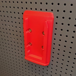 Puede incluir: Un soporte de pared de plástico rojo con cuatro agujeros para tornillos. El soporte está diseñado para sujetar un dispositivo u objeto.