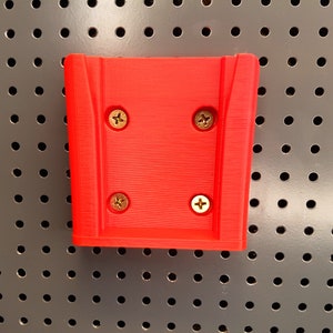 Puede incluir: Un soporte de pared de plástico rojo con cuatro tornillos dorados. El soporte tiene una superficie texturizada y está diseñado para sujetar un objeto.