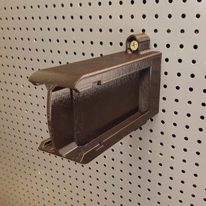 Puede incluir: Un soporte de pared de plástico negro con un tornillo en la esquina superior. El soporte está diseñado para sujetar un objeto pequeño, como un teléfono o una tableta.