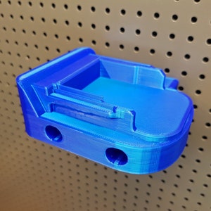Puede incluir: Un soporte de pared azul impreso en 3D para una batería. El soporte tiene dos agujeros para sujetarlo a una tabla de clavijas.