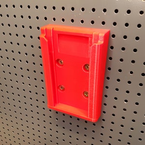 Puede incluir: Un soporte de pared de plástico rojo con cuatro agujeros para tornillos. El soporte está diseñado para sujetar un objeto pequeño, como un teléfono o una tableta.