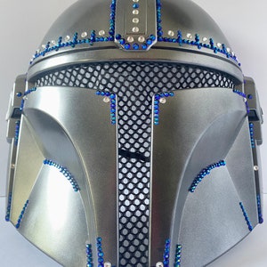 May include: A silver-colored helmet with a black mesh visor, adorned with blue and clear rhinestones. The helmet's design features a rounded top and angular facial features, with the rhinestones outlining the edges.