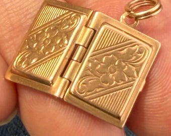 May include: A gold-toned metal charm shaped like an open book with a central hinge. Each rectangular cover features an embossed floral pattern set against a background of diagonal lines. A small circular jump ring is attached to the top corner.