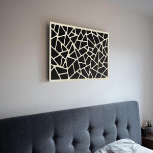 Puede incluir: Una obra de arte mural moderna con un diseño geométrico. La obra presenta un patrón negro de formas irregulares sobre un fondo crema. La pieza está montada en una pared sobre un cabecero tapizado gris.