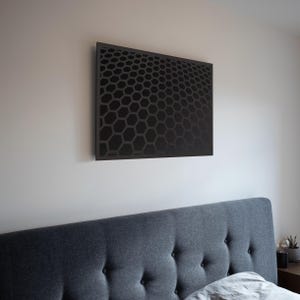 Peut inclure: Une œuvre d'art murale noire et géométrique avec un motif en nid d'abeille est accrochée sur un mur blanc au-dessus d'un lit rembourré gris. Le design de l'œuvre crée un effet tridimensionnel, ajoutant une touche moderne à la chambre.