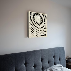 Puede incluir: Una pieza de arte mural cuadrada, de color crema, con un patrón de puntos negros dispuestos en líneas curvas. La obra de arte está montada en una pared gris claro, encima de un cabecero tapizado gris. Los puntos crean un efecto visual de onda.