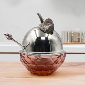 Puede incluir: Un mantequillero de vidrio rosa y plateado con forma de manzana con una pequeña cuchara de plata. La tapa tiene un diseño de hoja de plata.