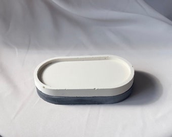 Puede incluir: Una bandeja ovalada de hormigón blanco con una base gris. La bandeja es perfecta para guardar objetos pequeños como joyas o llaves.