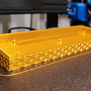 Puede incluir: Una carcasa de teclado impresa en 3D de color dorado con un patrón de panal en la parte inferior y los lados. La carcasa está sobre una superficie negra.