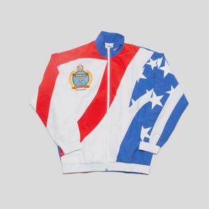 Op de afbeelding: Een wit, rood en blauw trainingsjack met ritssluiting en een patriottisch ontwerp. De jas heeft een wapen op de borst en een sterrenpatroon op de mouwen. De jas heeft een blauwe kraag en manchetten.