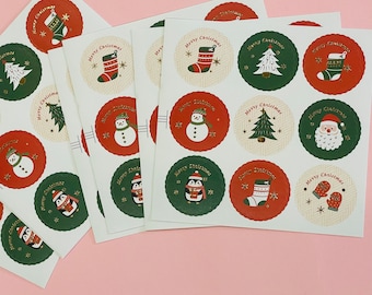 Puede incluir: Varias hojas de pegatinas con temática navideña. Cada pegatina circular presenta diseños festivos como muñecos de nieve, árboles de Navidad y calcetines, sobre fondos rojos, verdes y crema. Las palabras "Merry Christmas" están impresas en cada pegatina.
