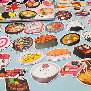 May include: A collection of colorful stickers featuring various Japanese foods, including sushi, ramen, rice, and other dishes. The stickers are arranged on a light blue surface.