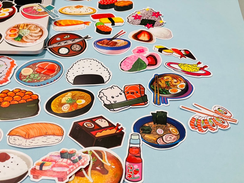 May include: A collection of colorful stickers featuring various Japanese food items, including sushi, ramen, and tempura. The stickers are arranged on a light blue surface.