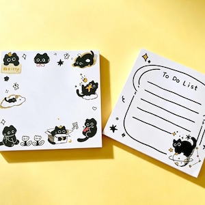 Puede incluir: Dos blocs de notas cuadrados de papel blanco con ilustraciones de gatos negros. El bloc izquierdo tiene un borde de gatos, estrellas y flores con el texto kitty. El bloc derecho tiene forma de rebanada de pan con el texto To Do List y líneas.