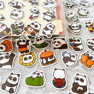 May include: A selection of panda bear stickers in different poses and activities. The stickers depict pandas with honey, on a scooter, with a burger, and other designs. The stickers are black, white, and tan with a white border.