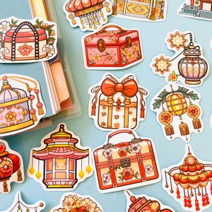 Può includere: Una collezione di adesivi colorati con vari disegni di ispirazione asiatica. Gli adesivi includono lanterne, scatole regalo e pagode, tutti adornati con elementi floreali e decorativi nei toni del rosso, rosa e oro. Gli adesivi sono su uno sfondo azzurro.