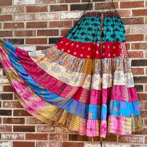 May include: A colorful, tiered skirt with a bohemian style. The skirt features a variety of patterned fabrics in shades of pink, blue, red, and gold. The skirt is hanging against a brick wall.