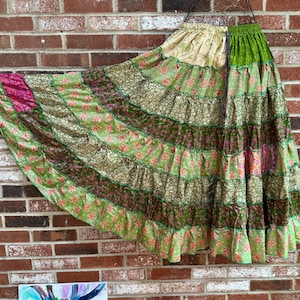 May include: A long, tiered skirt featuring a patchwork design of various green, cream, and magenta fabrics. The skirt has an elasticized waistband and is constructed from multiple horizontal panels with floral and geometric patterns throughout.