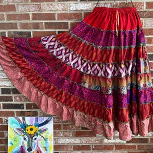 May include: A long, tiered skirt in shades of red, purple, and pink, with a drawstring waist. The skirt features various patterned fabrics, including paisley and geometric designs. The hemline has a ruffled edge. The skirt is hanging against a brick wall.