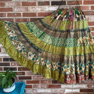 May include: A long, tiered skirt with a patchwork design. The skirt features various patterns and colors, including green, brown, and floral prints. It is hanging against a brick wall. The skirt has a drawstring waist and a flowing silhouette.