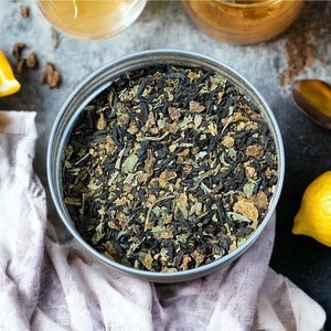 Pu&ograve; includere: Un contenitore di metallo argentato pieno di miscela di t&egrave; sfuso. La miscela di t&egrave; &egrave; un mix di t&egrave; nero, erbe e spezie. Il contenitore &egrave; su una superficie grigia con limoni e altri ingredienti.