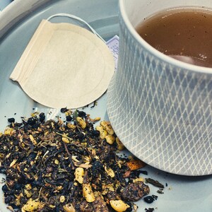 May include: A close-up of a light blue ceramic mug filled with a dark brown tea. A tea bag sits on a light blue saucer with loose tea leaves scattered around it.