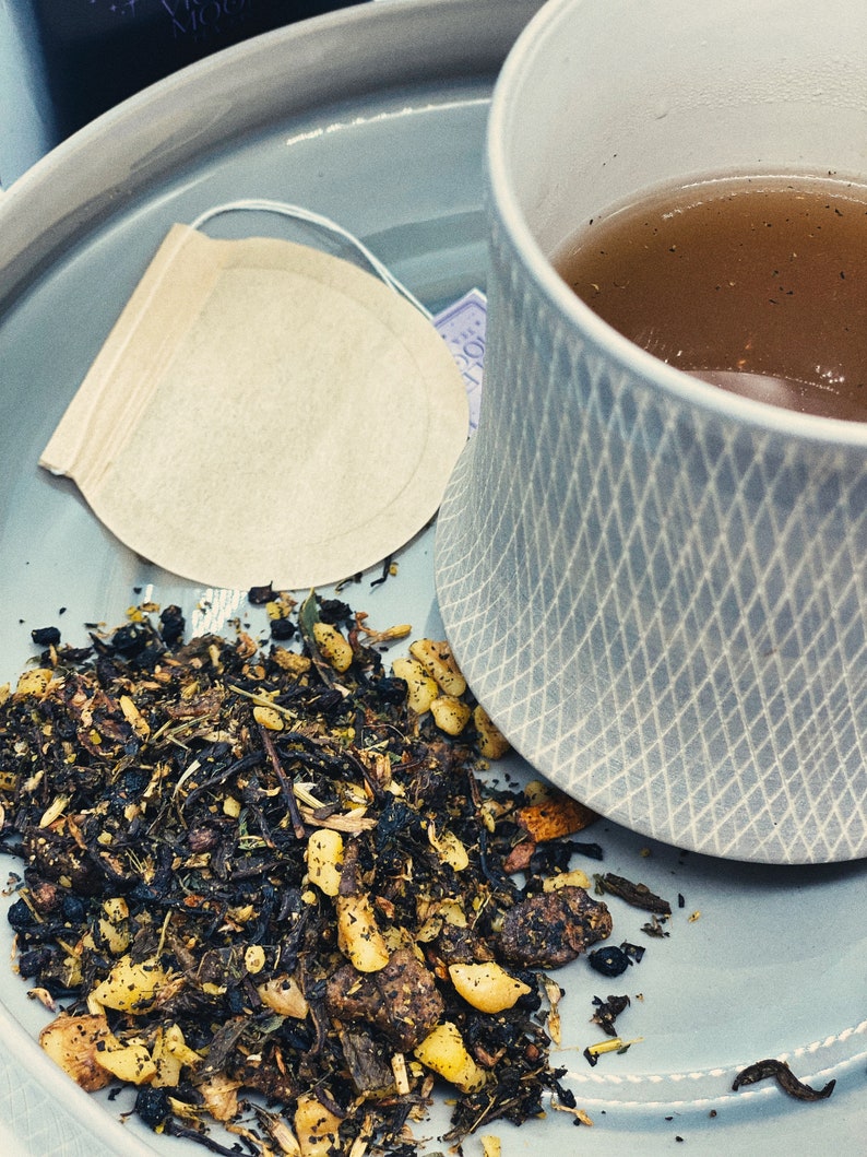 May include: A close-up of a light blue ceramic mug filled with a dark brown tea. A tea bag sits on a light blue saucer with loose tea leaves scattered around it.