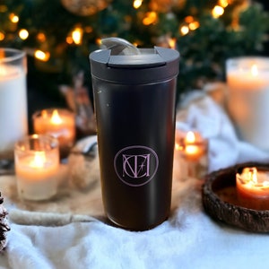 May include: A black travel mug with a lid and a pink monogram "NZ" on the front. The mug is sitting on a white surface with candles and a pine cone.