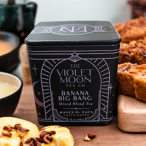 May include: A black tin container of loose leaf tea with a purple and white label. The label reads "The Violet Moon Tea Co. Banana Big Bang Mixed Blend Tea Makes 15+ Cups Caffeinated 1.6 oz (46g)"