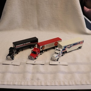 May include: Three miniature semi-trucks labeled 1, 2, and 3. Truck 1 is black with a Midnight X-Press trailer. Truck 2 is red with a Coca-Cola trailer. Truck 3 is white with a blue and red striped trailer. Each truck has a cab and a trailer.