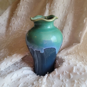 May include: A ceramic vase with a ruffled rim and a gradient of colors. The top is a seafoam green, transitioning to a light blue, and then a deep navy blue at the base. The vase has a glossy finish.