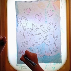 Op de afbeelding: Een kind kleurt een tekening van een eenhoorn met een lichtblauw lichaam en een roze manen op een lichttafel. De tekening bevat ook twee bloemen en drie harten. De lichttafel is geel en heeft de tekst "Beezy Baby Bees" onderaan.
