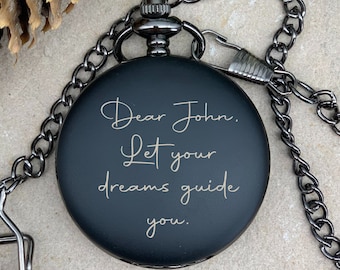 May include: Black pocket watch with a silver chain. The watch face has an engraved message that reads "Dear John. Let your dreams guide you."