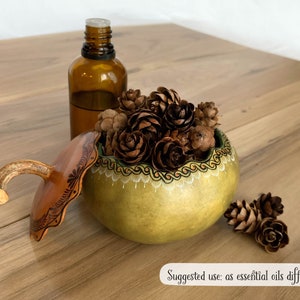 May include: A handcrafted gourd-shaped essential oil diffuser with a light green body and a decorative rim. The lid is brown with etched designs. The diffuser is filled with pine cones, and a brown glass bottle is next to it.