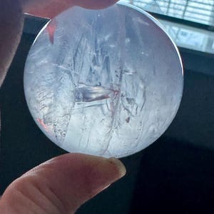 May include: A clear, round crystal ball held between fingers. The crystal has internal fractures and inclusions, with a slightly cloudy appearance. The light refracts through the sphere, creating interesting patterns.
