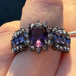 May include: A black ring with a large purple gemstone surrounded by smaller clear stones. The ring has a detailed design with black metal accents.