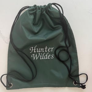 May include: A dark green drawstring bag with black drawstrings. The bag has the embroidered text "Hunter Wildes" in white, elegant script. The bag is made of a smooth, possibly waterproof material.