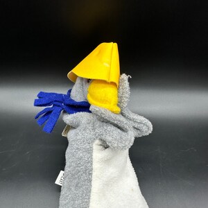 May include: Gray and white puppet with a yellow hat and blue scarf. The puppet is wearing a yellow hat with a pointed top and a blue scarf around its neck. The puppet is made of soft, fuzzy material.